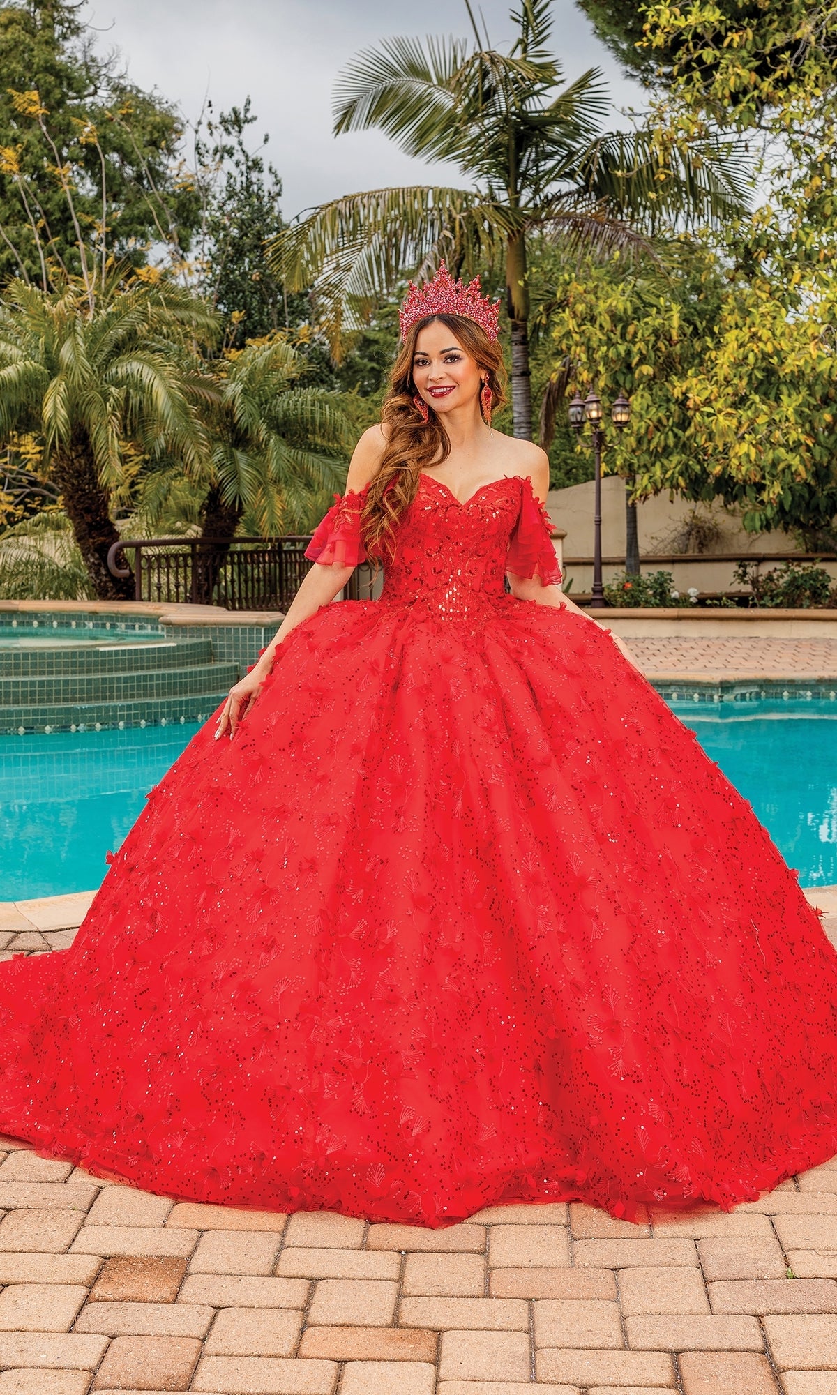 A young woman wearing the Quinceanera Dress 1849 by Dancing Queen, adorned and crowned to match, stands by a pool with palm trees in the background.